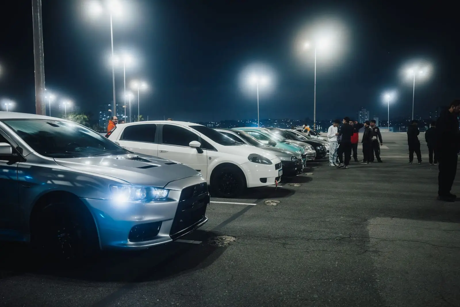 Cars and people gathered under floodlights at a night-time car meet.