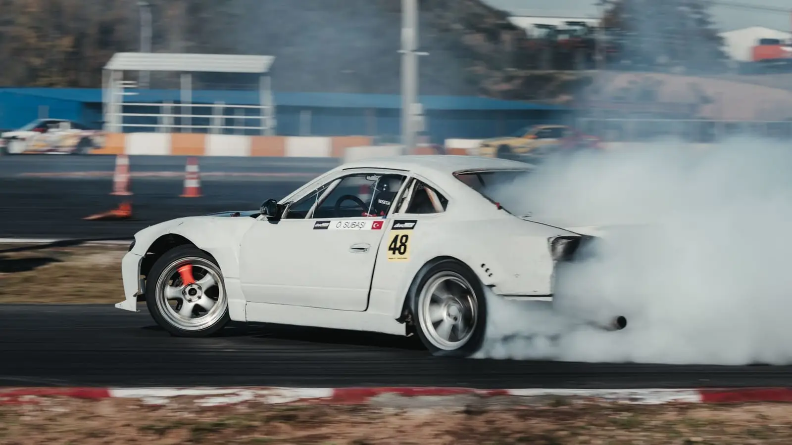 A white drift car slides sideways in a cloud of tire smoke.