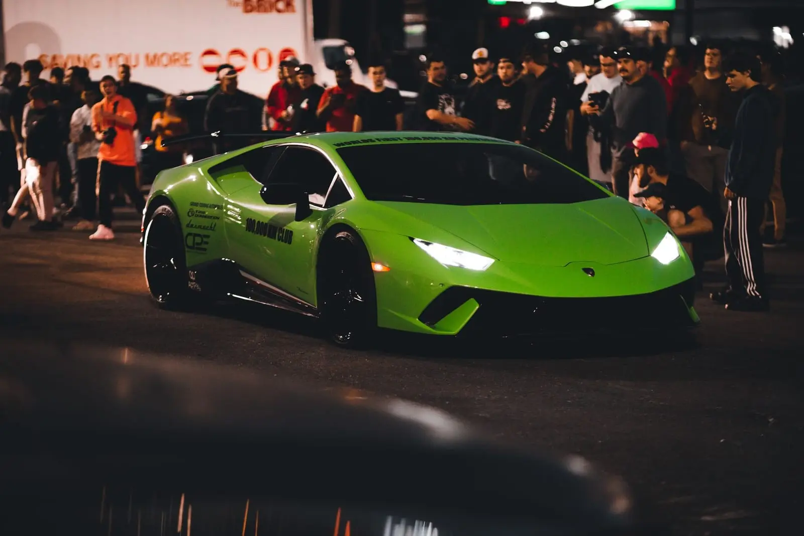 A green supercar rolls through a night-time car meet as the crowd looks on.