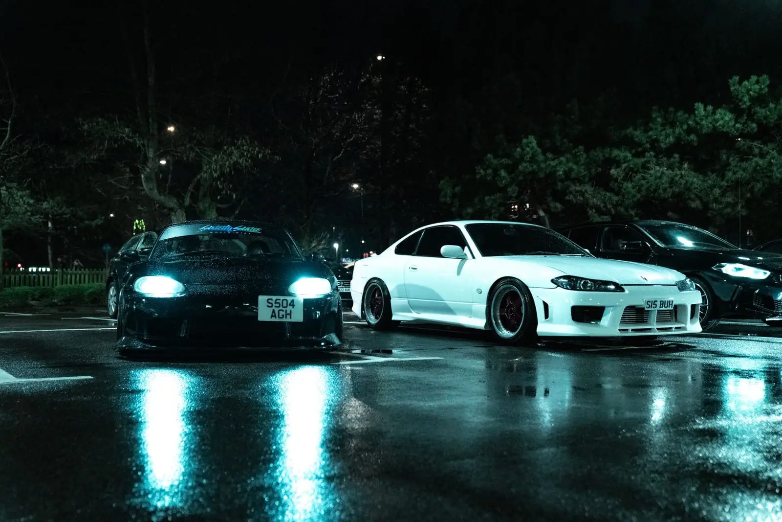 Classic Japanese sports cars reflected on a rain-soaked parking lot at night.