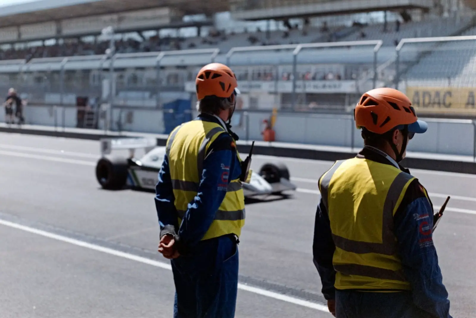 Two track marshals in orange helmets watch a single-seater pass on the pit straight.