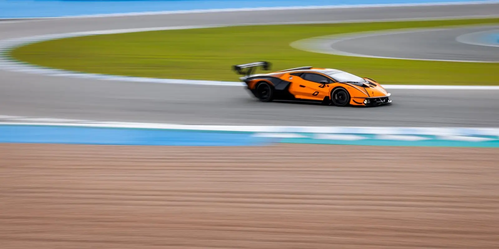 An orange GT race car leans through a sweeping corner on a sunny circuit.