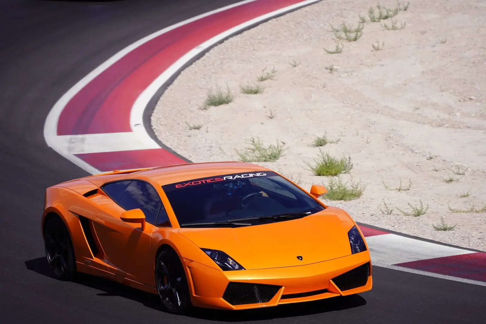 An orange supercar rounds a red and white kerb at a desert track.