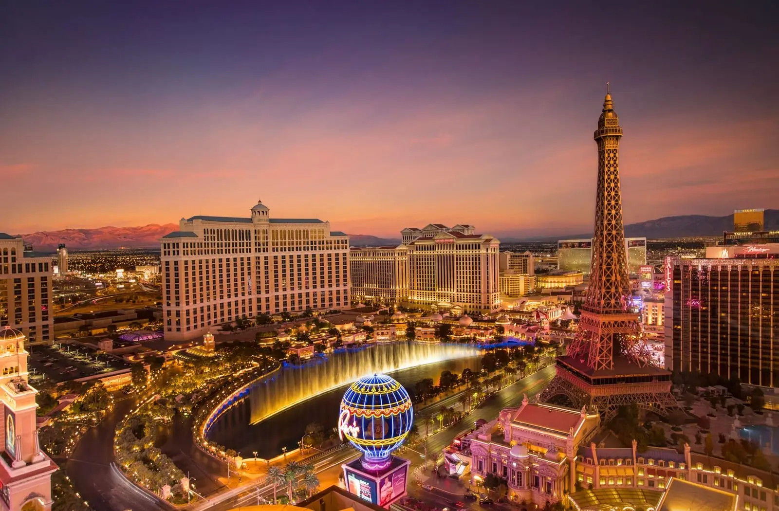 The Strip at dusk from above: Bellagio, the Eiffel Tower replica and the Paris balloon sign under a purple sky.