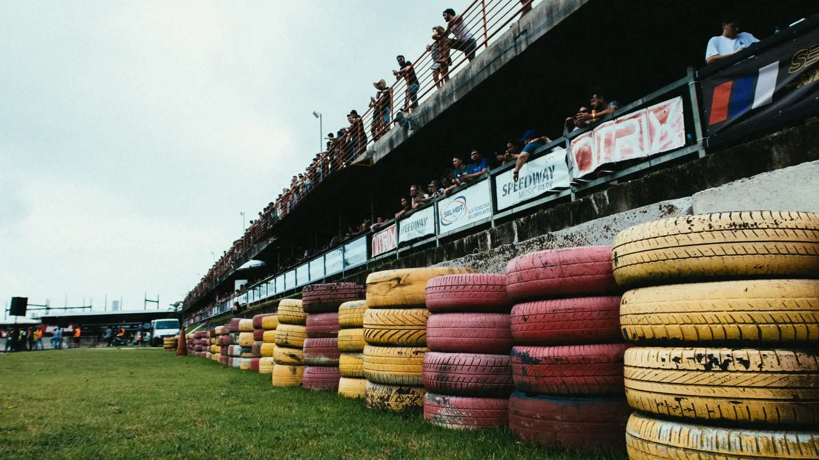 Spectators lean over the fence above a colourful tire wall at a race track.