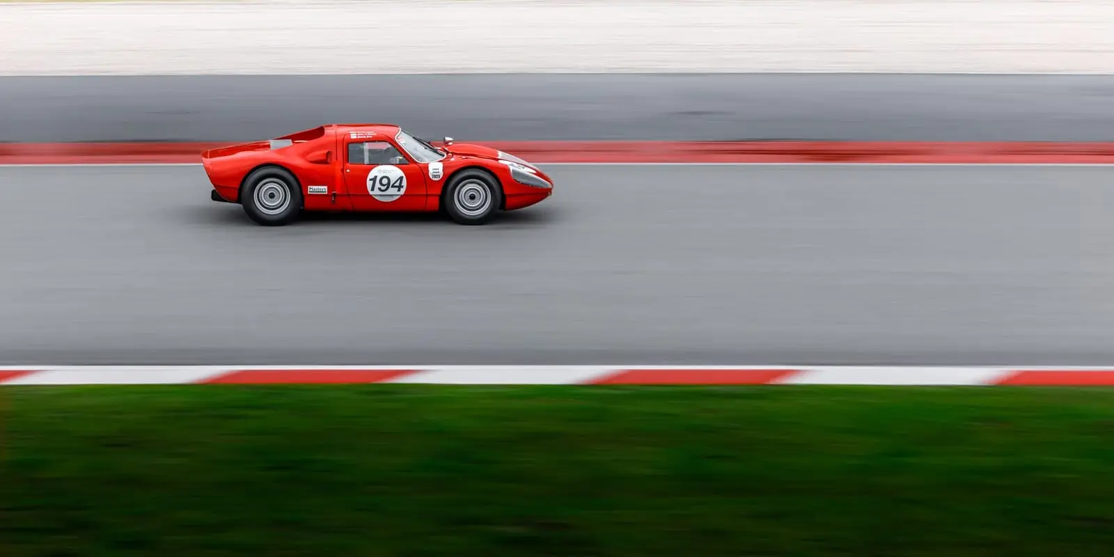 A vintage red race car blurred with speed along a straight lined with red and white kerbs.
