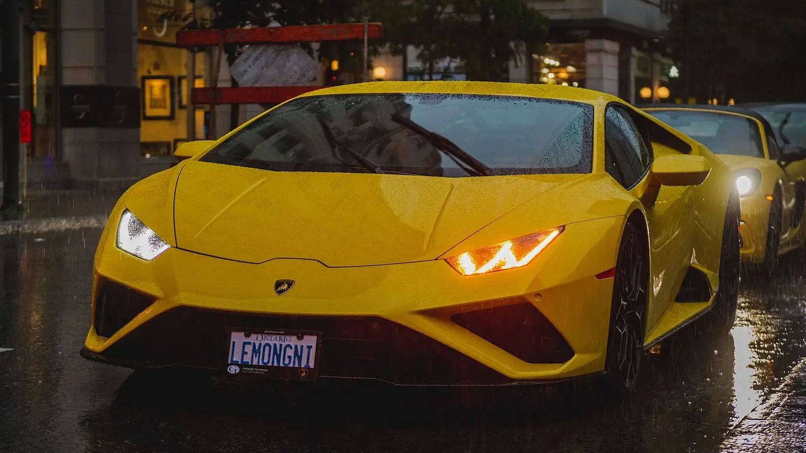 A yellow supercar with its lights on, on a wet street at night.